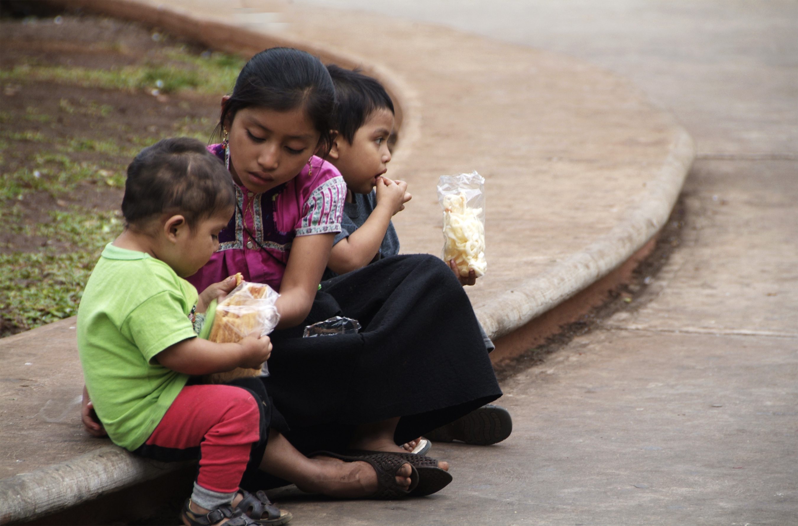Three kids sit on a curb in Mexico while eating a snack. Margie Nea for Bread for the World.